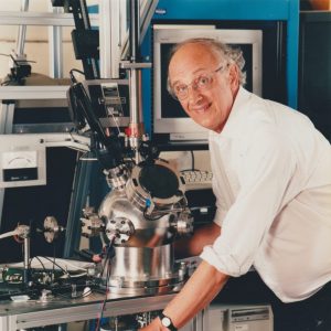 Sir John Enderby leans over some scientific equipment and smiles warmly at the camera. He is an older white man with glasses, and looks happy and excited.